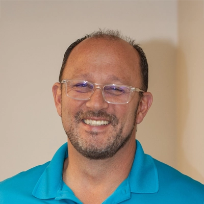 A close-up portrait of Dr. Steve Manuel wearing a teal collared shirt and clear-framed glasses