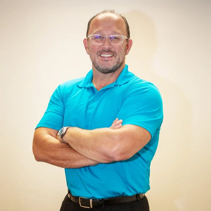 Dr. Steve Manuel standing against a plain, light-colored background, wearing a bright teal short-sleeve polo shirt